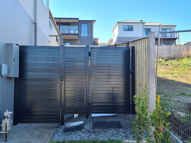 Black horizontal slatted driveway gates open to a gravel entrance with modern houses and fencing visible in background