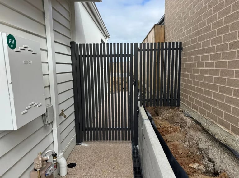 Narrow side yard with black metal vertical slat gate, brick wall, gravel ground, and white house siding