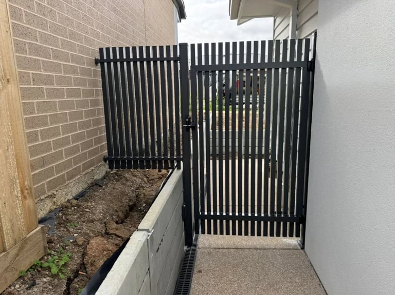 Black vertical slat gate and fence in narrow side yard with concrete path and brick wall