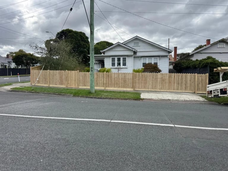 Single-story beige house with wooden fence on residential street under cloudy sky