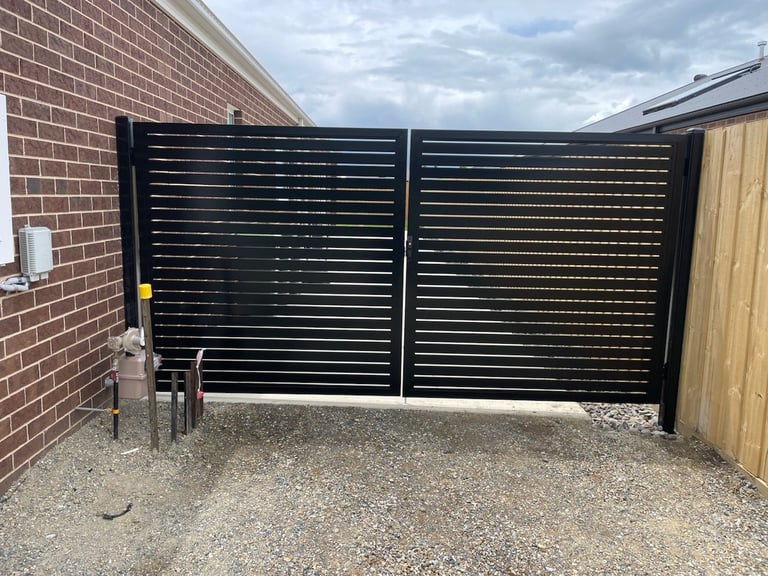 Black metal slatted driveway gates with horizontal bars mounted on a brick wall beside a wooden gate and gravel surface.