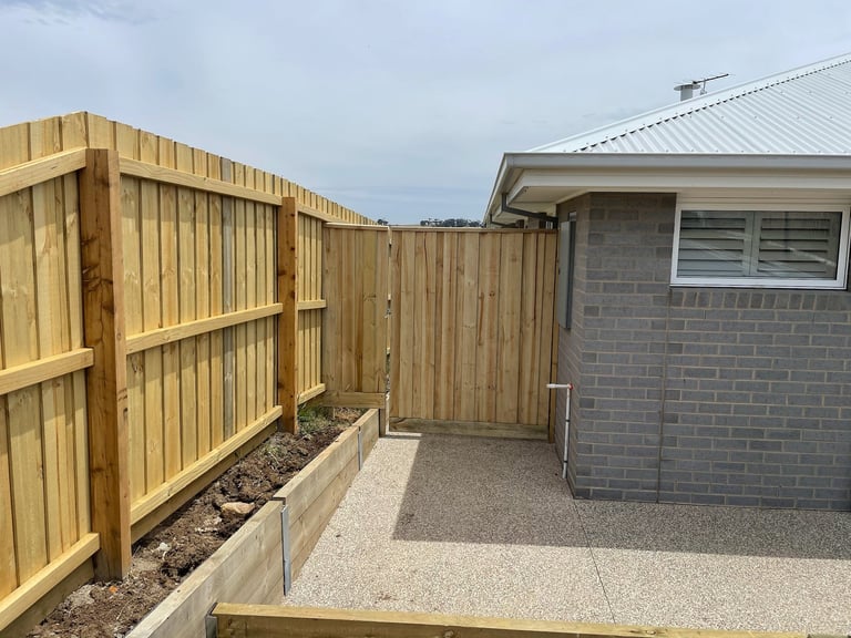 Backyard patio with wooden fence gate, gravel pathway, and modern gray brick building structure